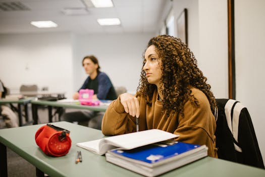 Young woman in classroom, focused on study material with peers in the background.